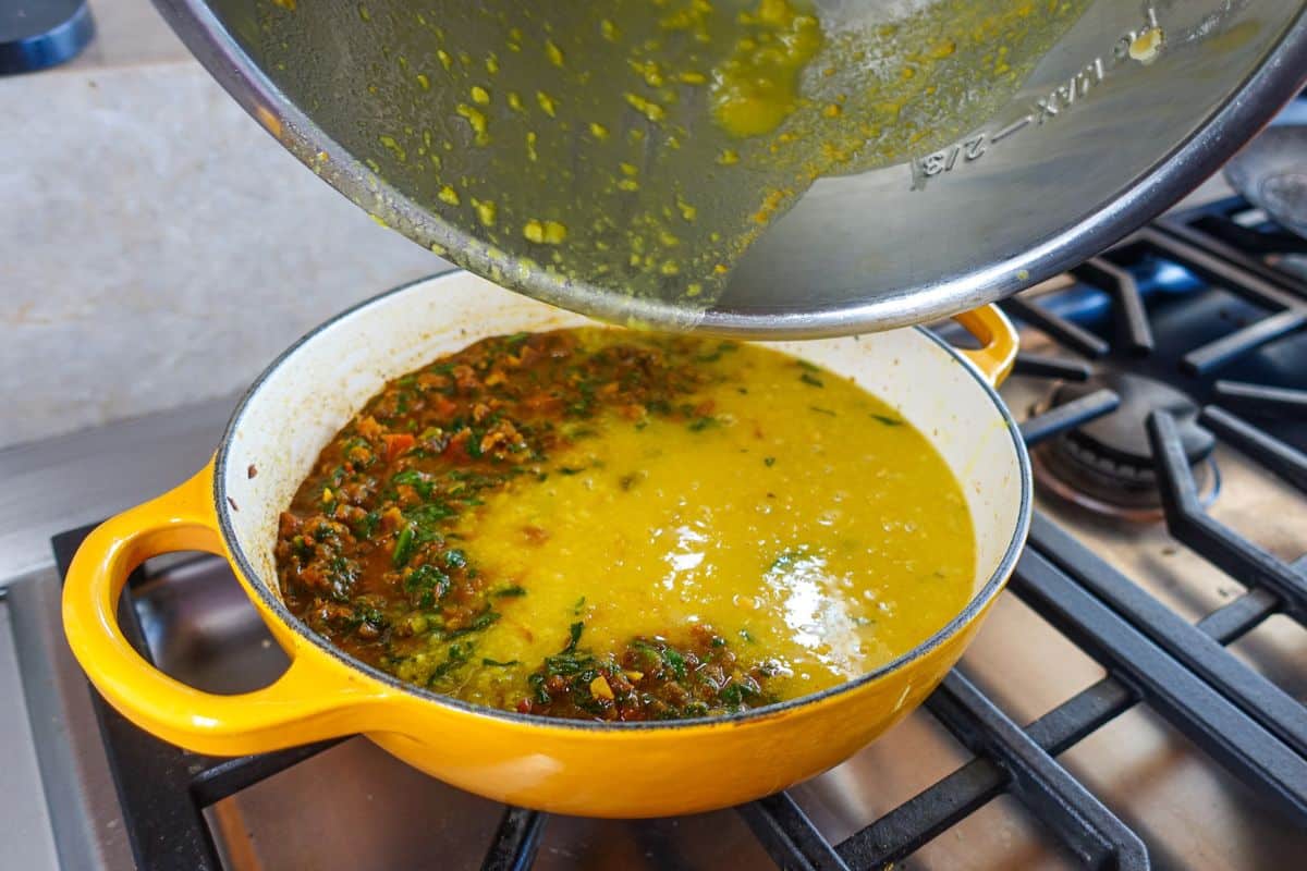 A pot of yellow dal with herbs cooks on a gas stove, while a lid is lifted to reveal the contents.