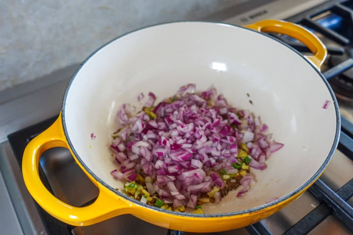 Chopped red onions are added in a yellow enameled pot on a stovetop.