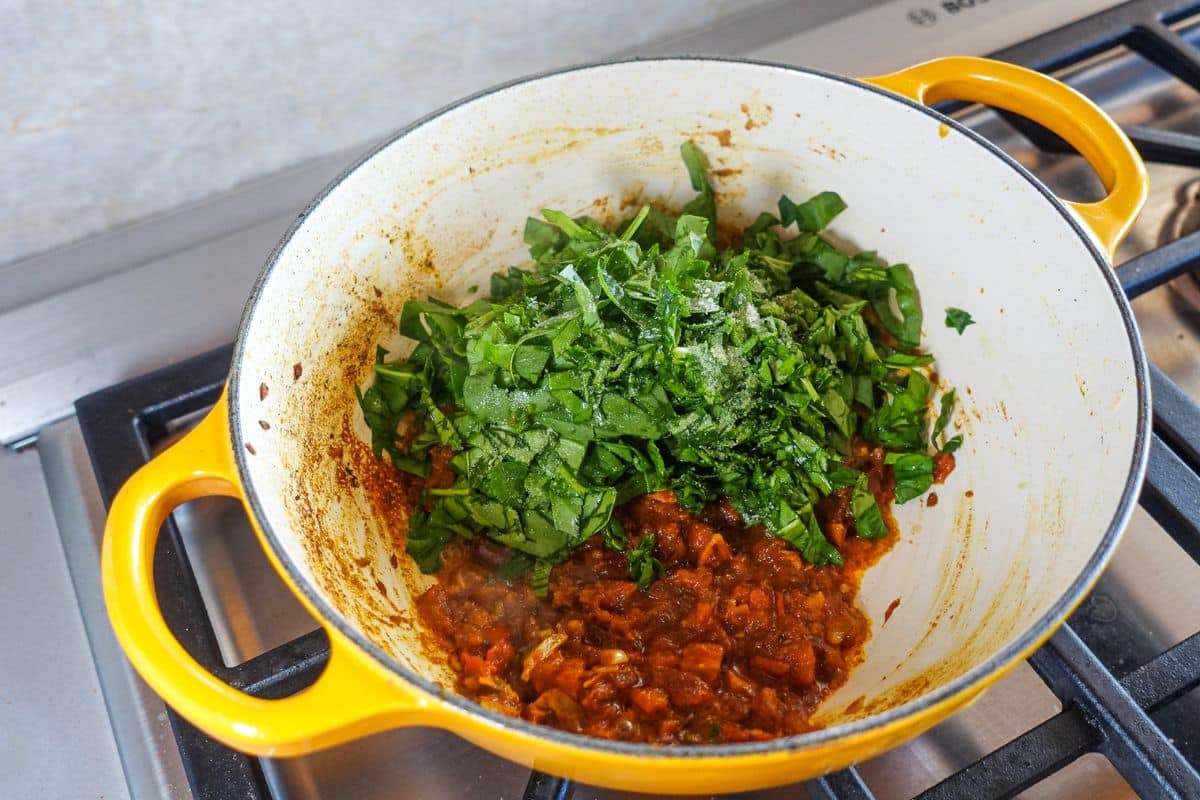 A yellow pot on a stove contains a mixture of chopped spinach and tomato-based masala.