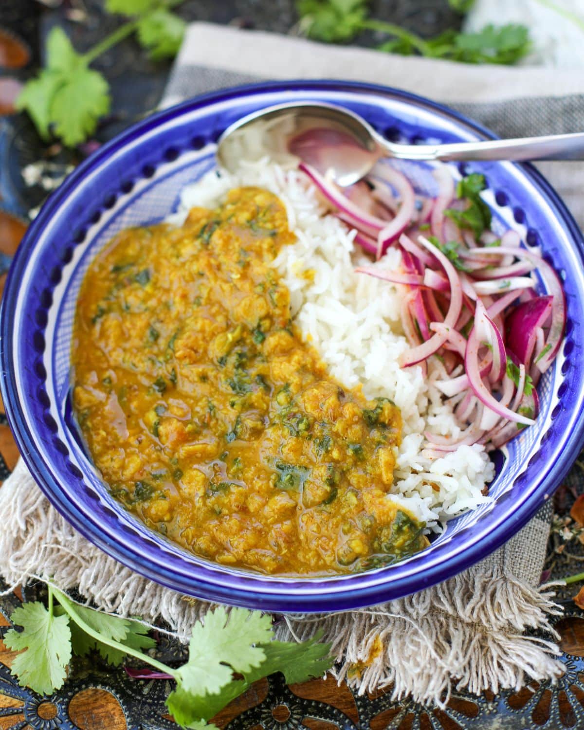 A blue bowl with spinach dal fry, white basmati rice, and sliced red onion salad, served with a spoon on a fringed cloth.