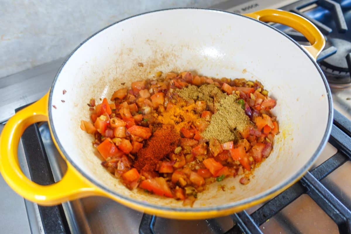 A yellow pot on a stove contains chopped tomatoes, onions, and assorted ground spices, being cooked together.