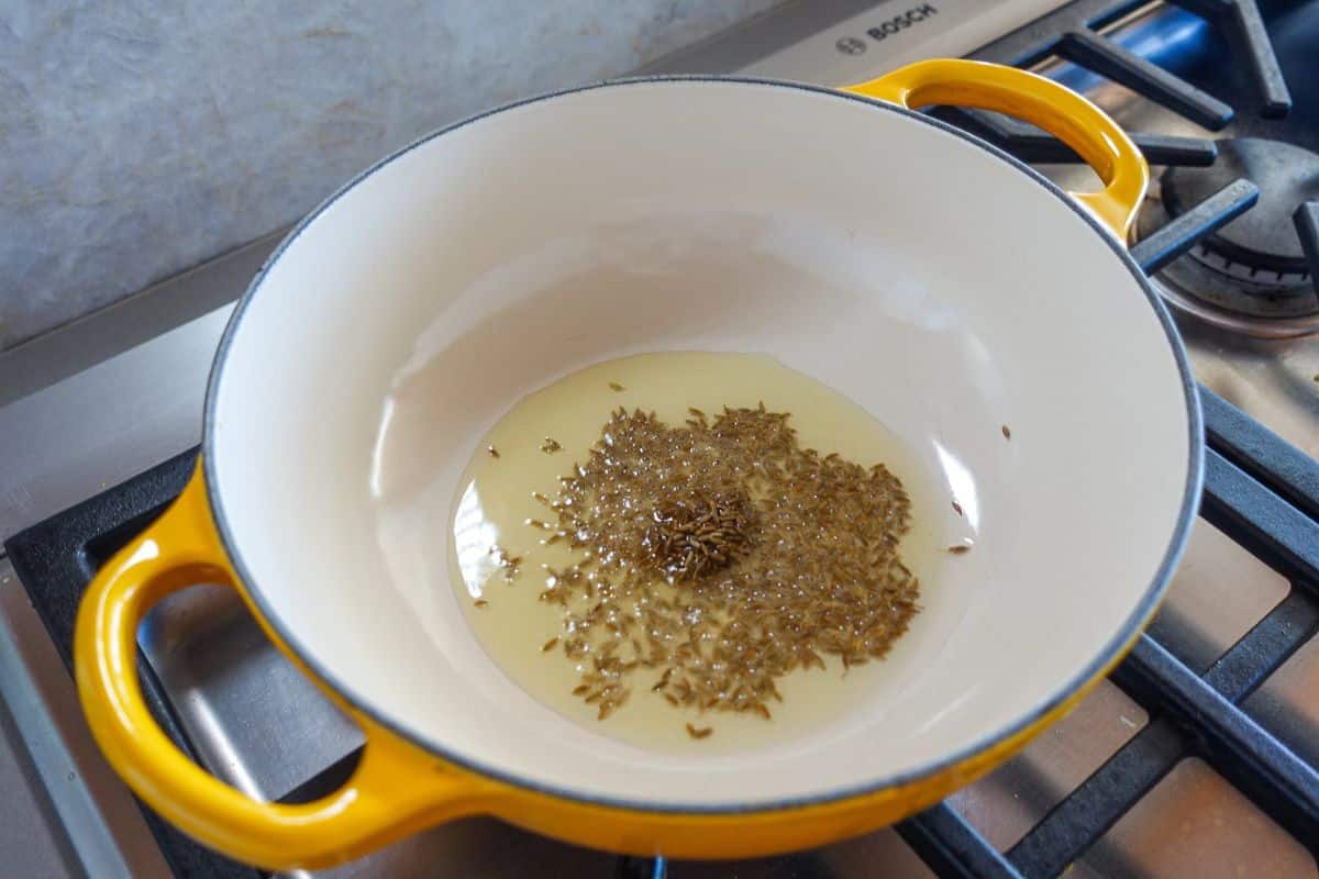A yellow pot on a stove contains hot oil with cumin seeds being sautéed.