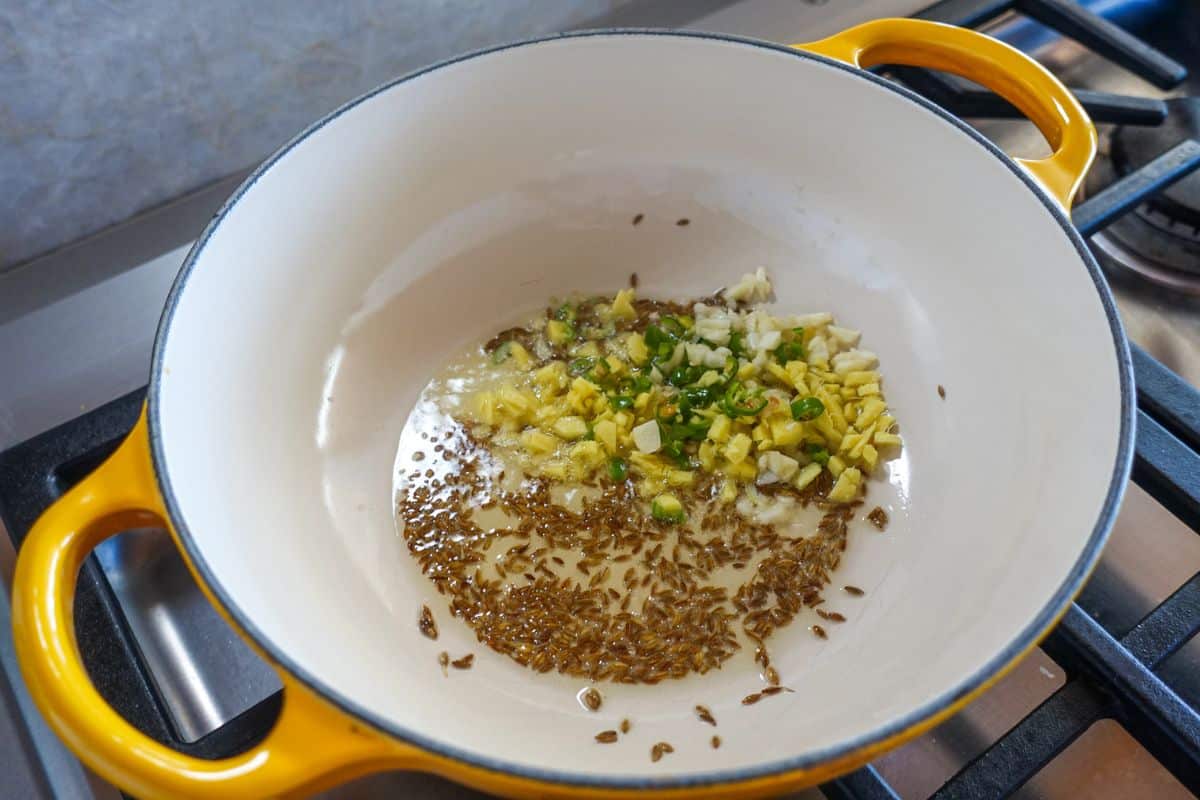 Chopped ginger, garlic, green chilies, and cumin seeds are being sautéed in oil in a yellow-handled pot on a stovetop.