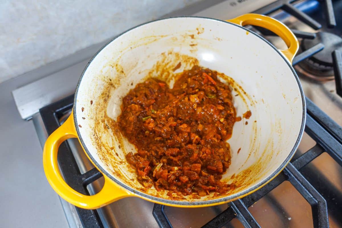Yellow Dutch oven on a stove containing cooked masala consisting of onions, tomatoes, ginger, garlic, and green chilies with spices.
