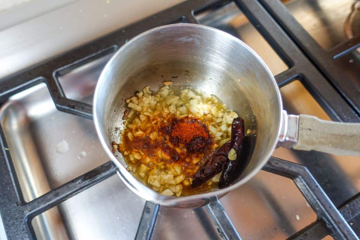 A saucepan on a stove with chopped garlic, whole dried chili peppers, oil, and Kashmiri red chili powder being sautéed for spinach dal fry.