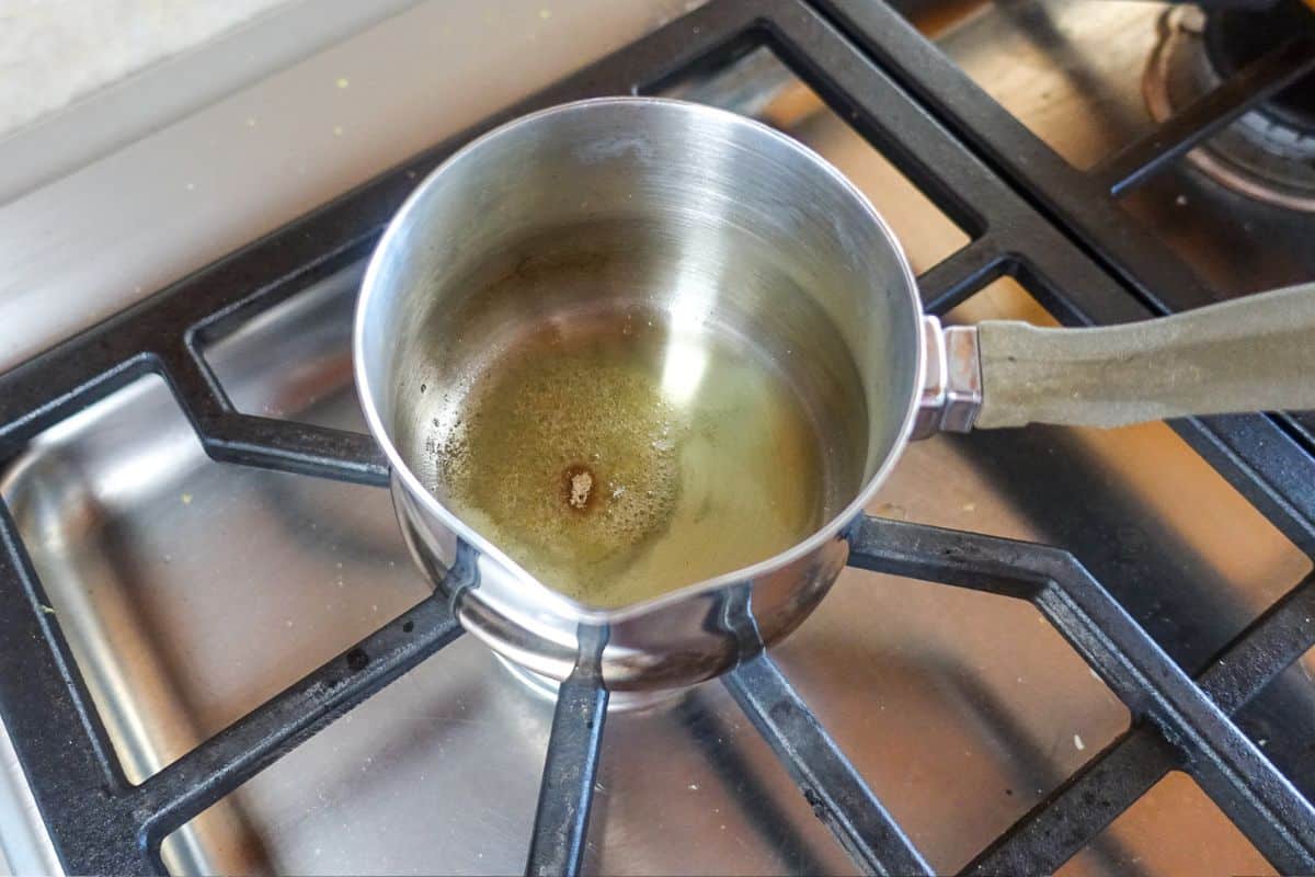 A small stainless steel pot with oil and asafoetida (hing) sits on a gas stovetop burner.