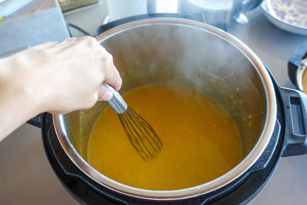 A hand uses a whisk to stir a pot of steaming yellow lentils inside an electric pressure Instant Pot on a kitchen counter.