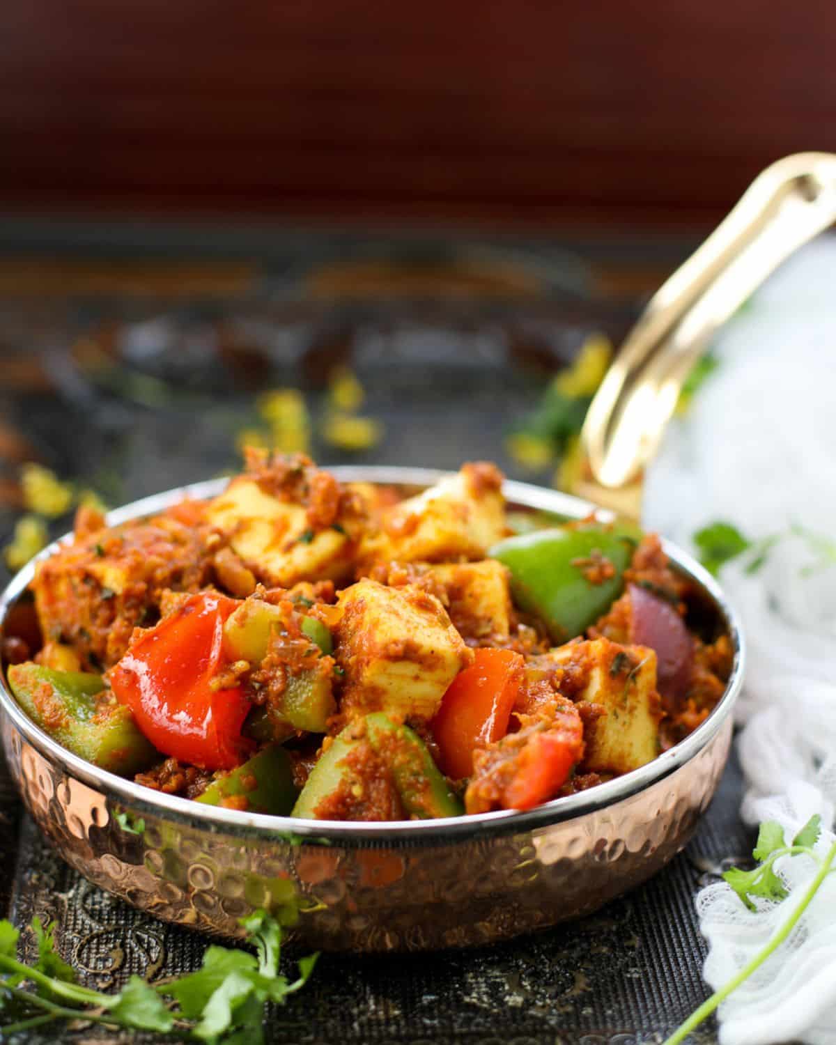 A copper bowl filled with kadai paneer, containing cubes of paneer, bell peppers, onions, and spices, placed on a dark surface.