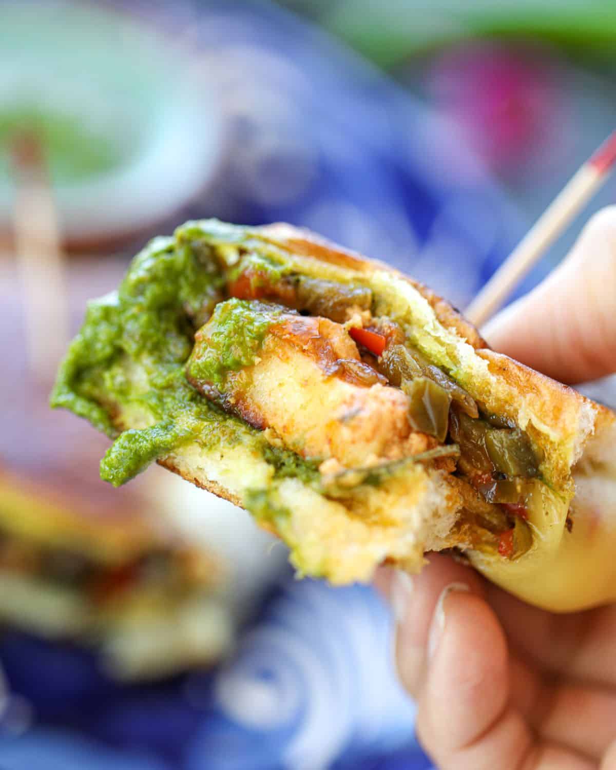 A hand holding a bite of the paneer tikka slider dipped in green chutney.