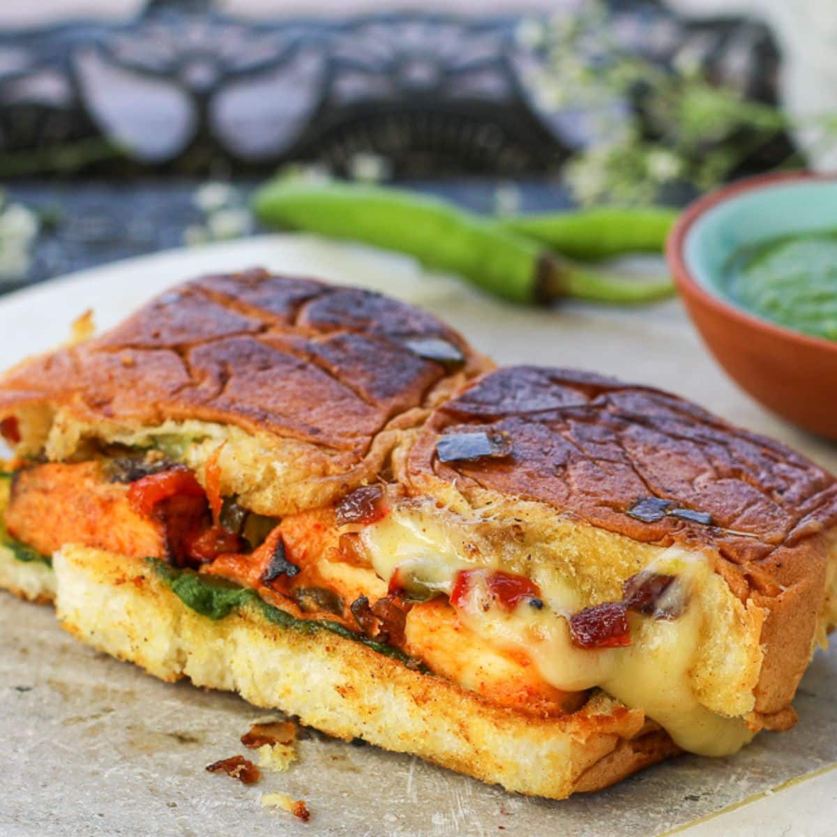Paneer Tikka Sliders with chutney, paneer, veggies, and cheese ozzing out with green chilies & chutney in the background.