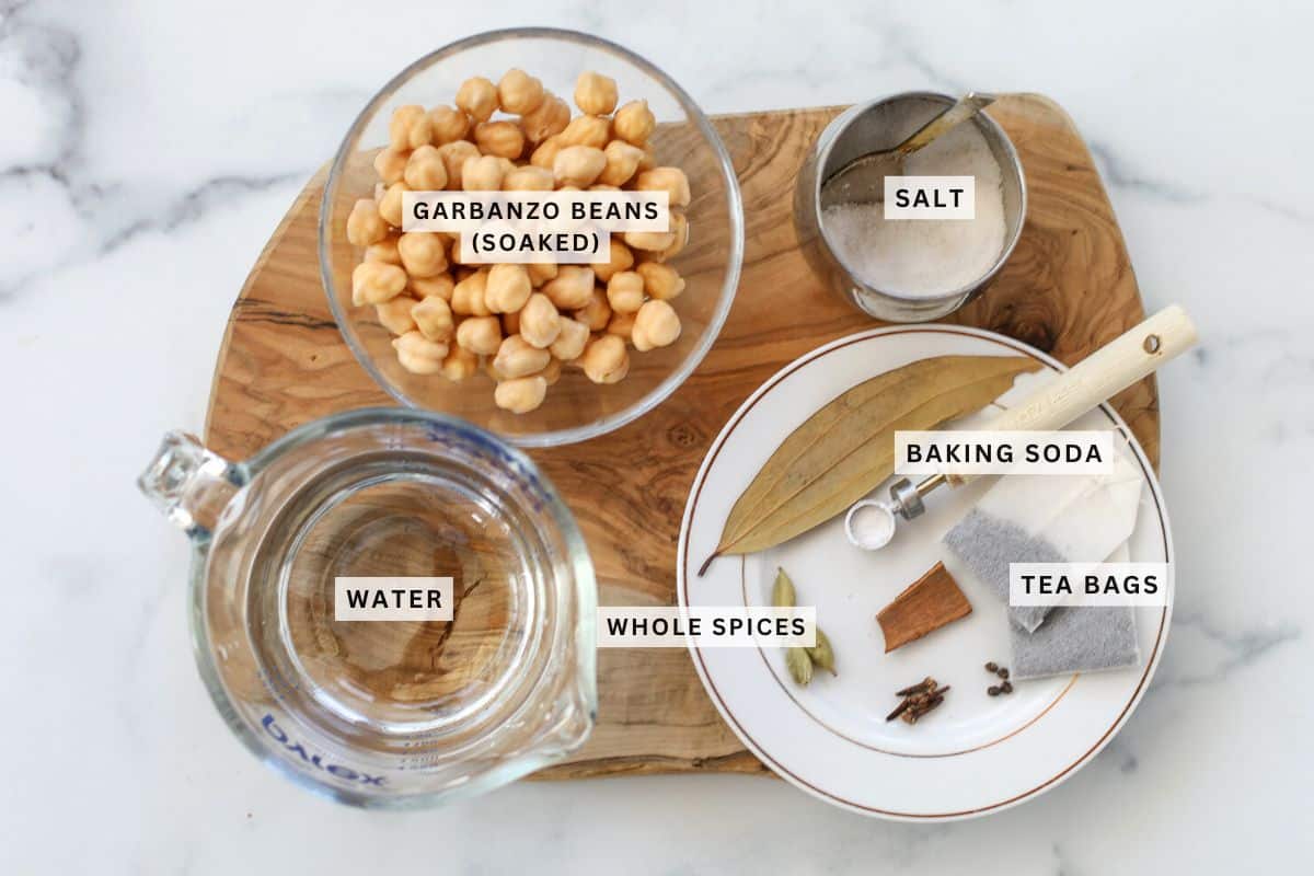 A wooden board holds bowls of soaked garbanzo beans and salt, a jug of water, whole spices, and tea bags. Each item is labeled.