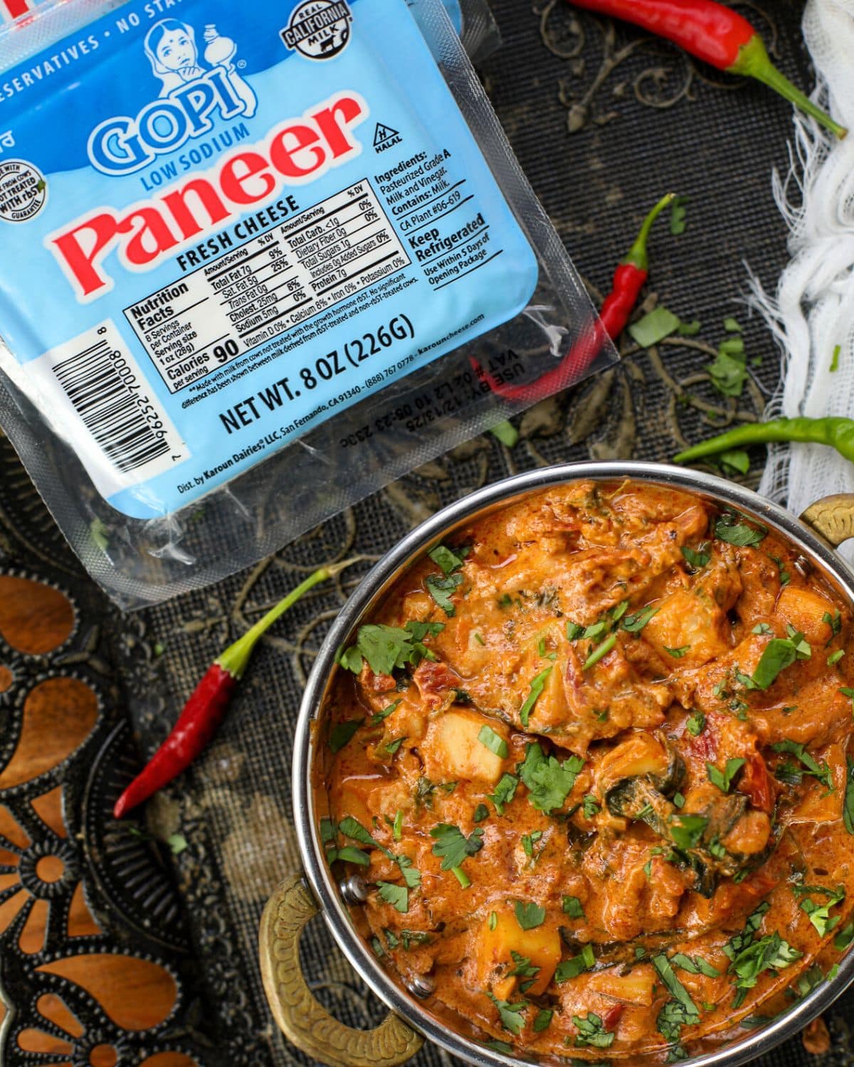 A copper bowl containing marry me paneer garnished with fresh cilantro with red & green chilies surrounding it and a package of Gopi paneer to the top left corner.