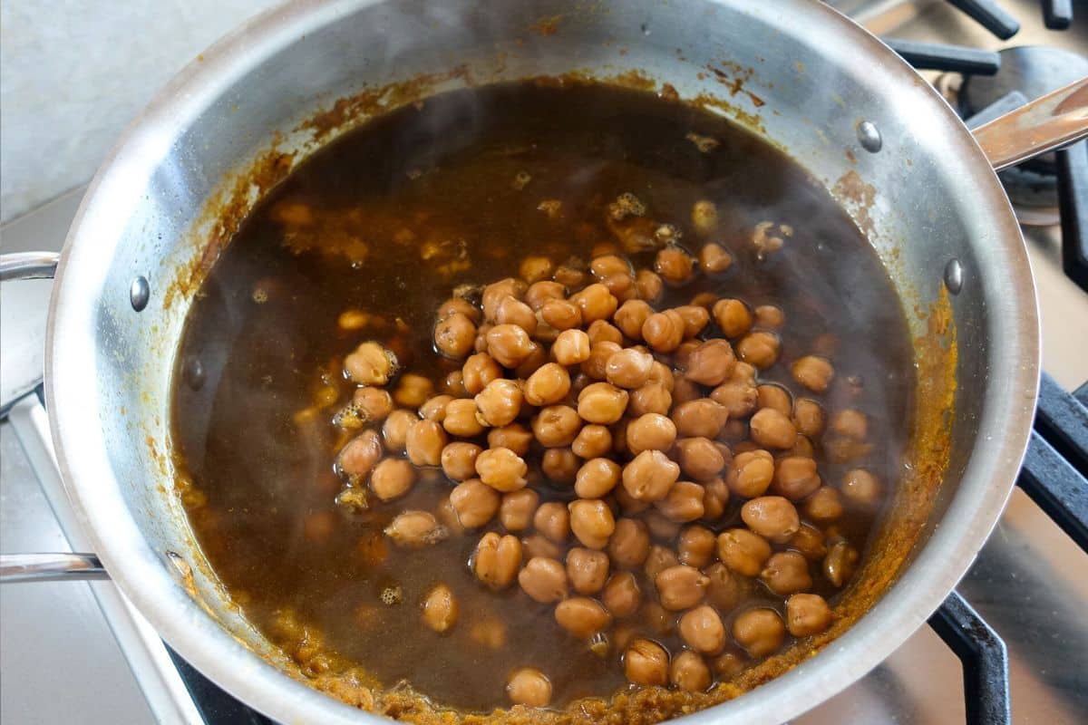 A stainless steel pan on a stove contains simmering chickpeas in a dark, seasoned liquid for Punjabi Chole. Steam is rising from the mixture.