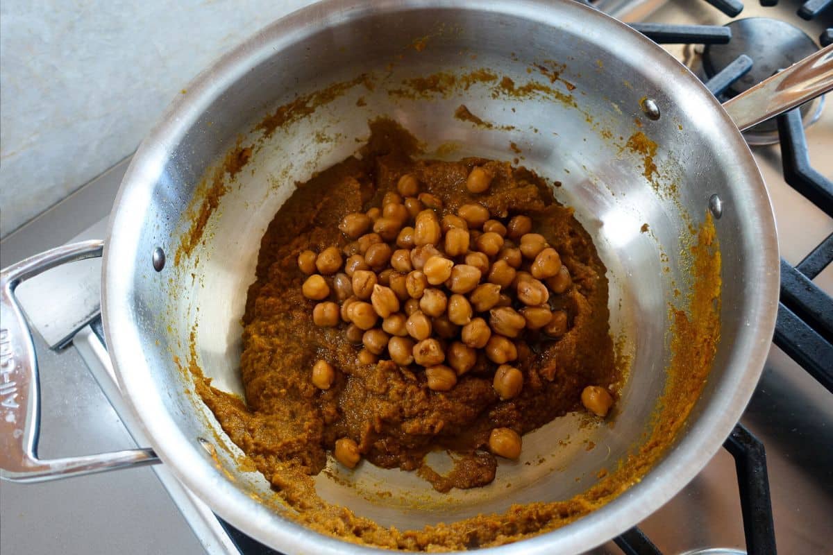 A metal pot on a stovetop contains a thick brown masala with whole chickpeas placed on top, ready to be mixed.