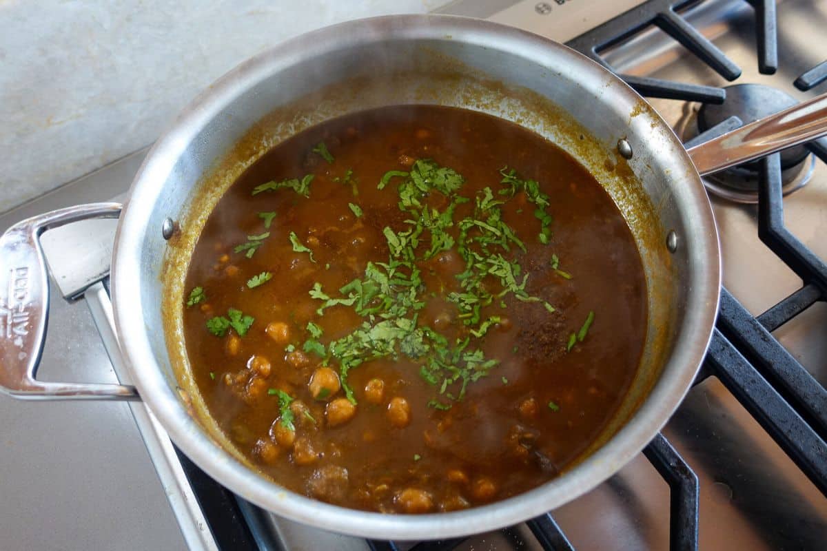 A stainless steel pan on a stove contains a brown chickpea curry garnished with chopped cilantro. Steam is rising from the dish.