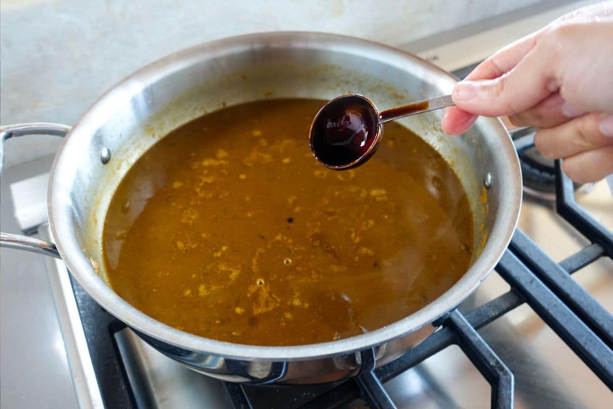 A hand holds a measuring spoon of tamarind concentrate above a saucepan filled with simmering brown chole on a stove.