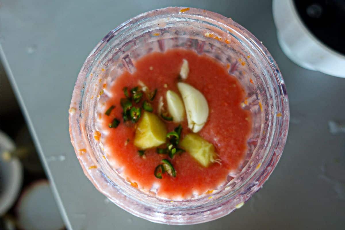 A blender containing a tomato puree mixture with visible garlic cloves, chopped green chili, and ginger pieces.