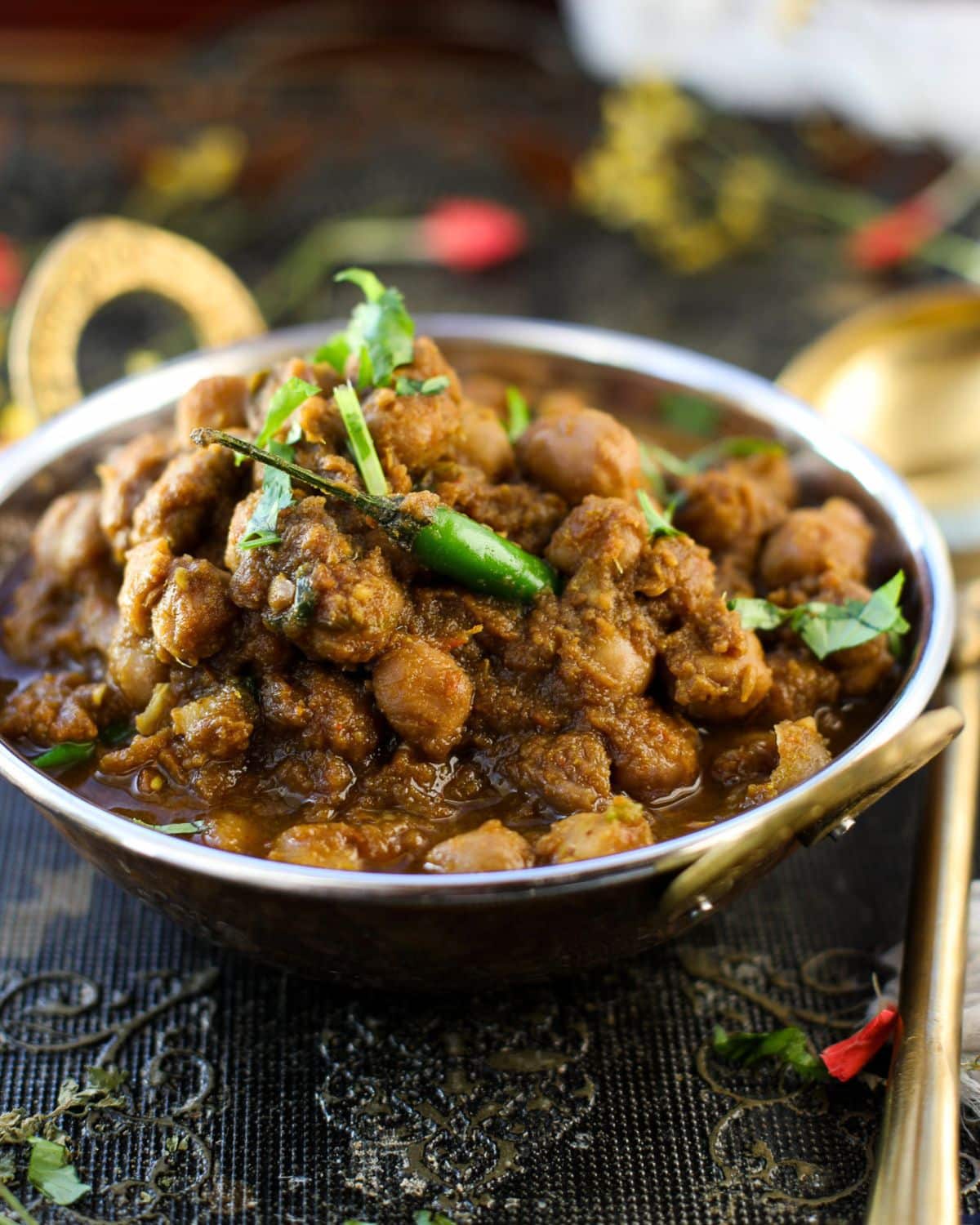 A copper bowl filled with Punjabi Chole garnished with cilantro and green chili, placed on a decorative dark tablecloth.