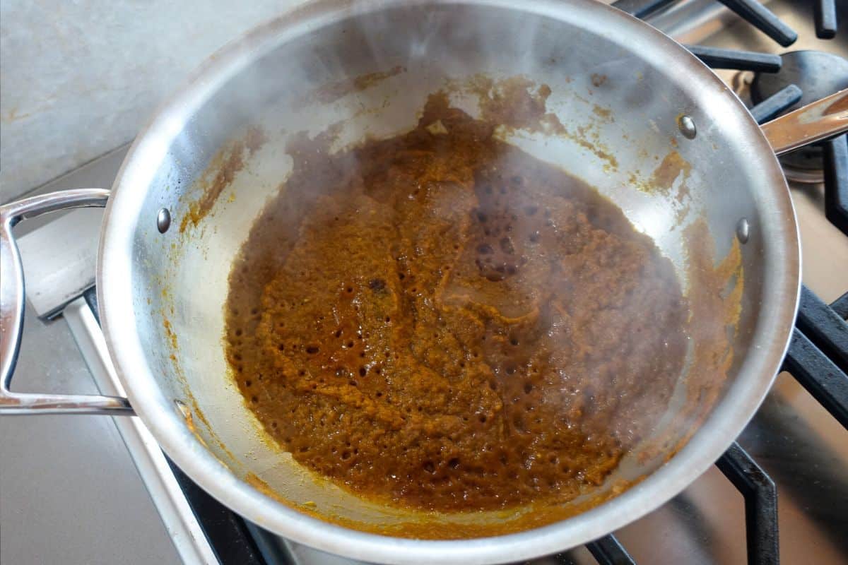 A stainless steel pan on a stovetop contains a brown masala, bubbling sauce with steam rising from it.