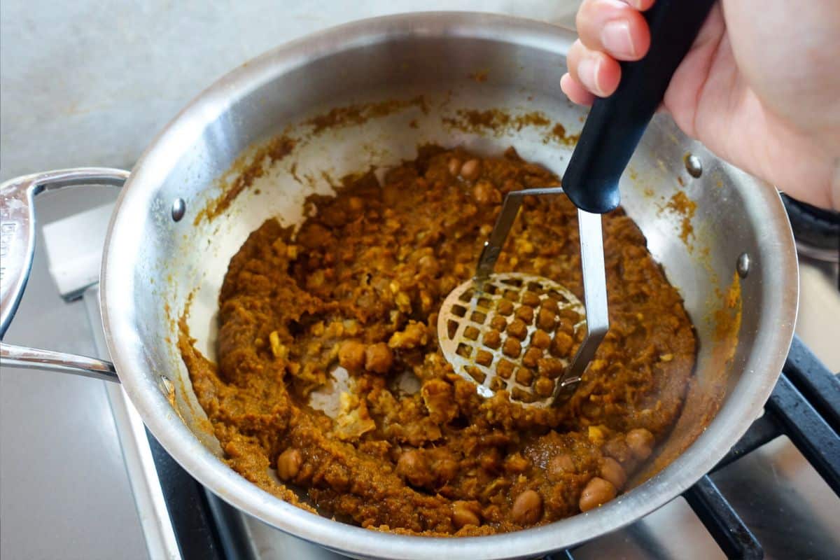 A hand uses a potato masher to mash chickpeas and spices in a metal pan on a stovetop.