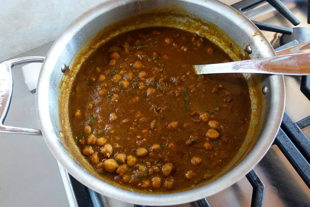 A stainless steel pan filled with Punjabi Chole sits on a stove, with a metal spoon resting inside the pan.
