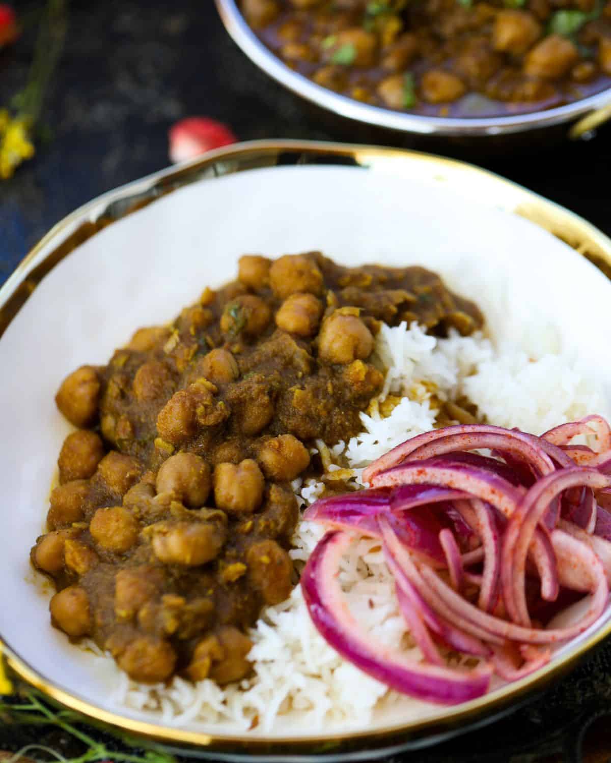 A bowl of white rice topped with Punjabi Chole and sliced red onions, with another bowl of curry in the background.