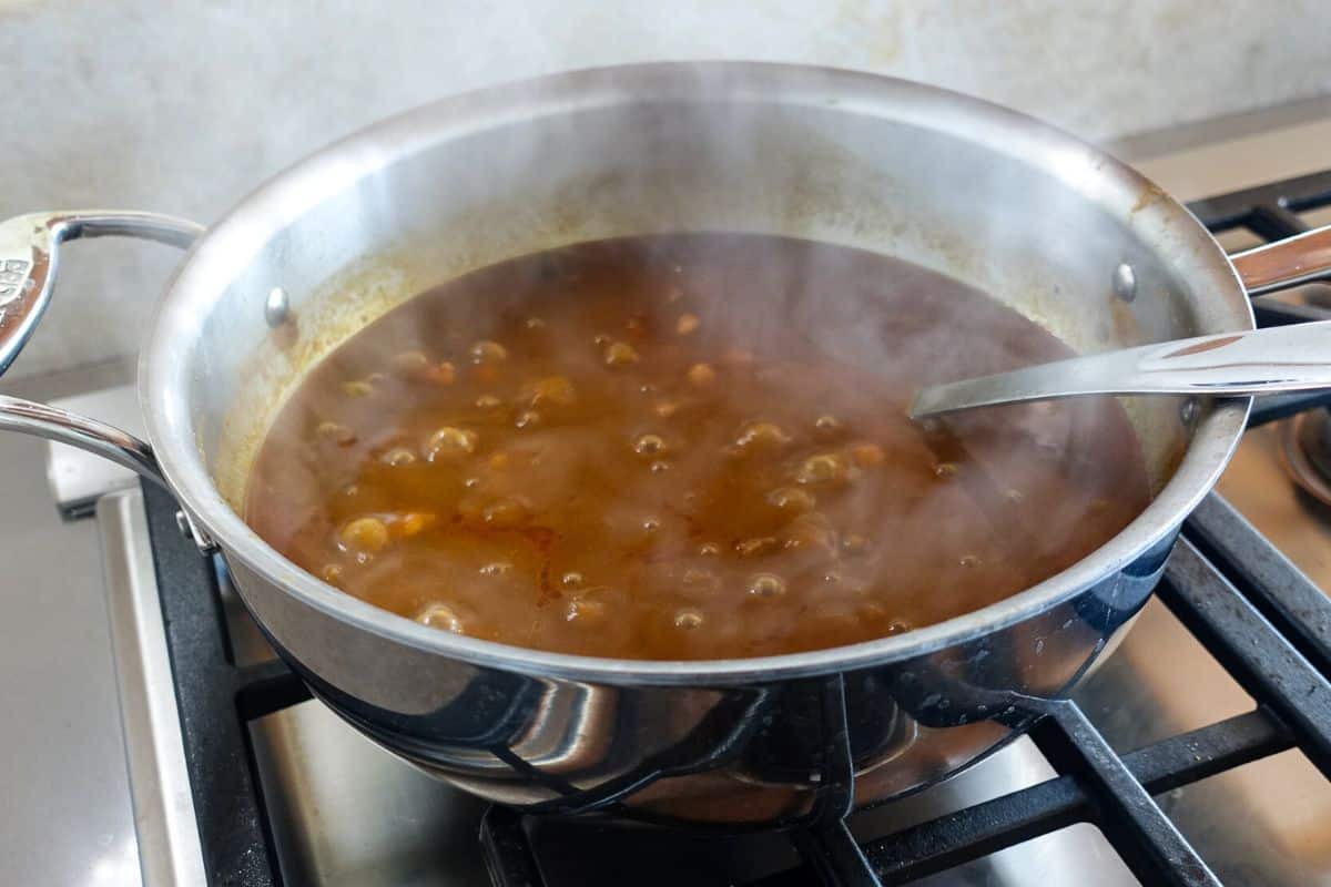 A stainless steel pot with a metal ladle sits on a stove, filled with bubbling brown Punjabi Chole, with steam rising from the surface.