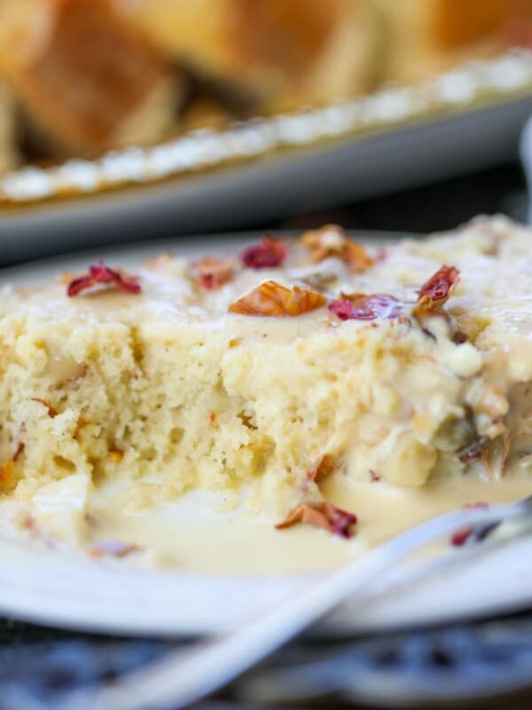 A slice of moist Ras Malai Nut Bread with nuts and glaze sits on a plate, a forkful missing; more cake pieces temptingly rest on a tray in the background.