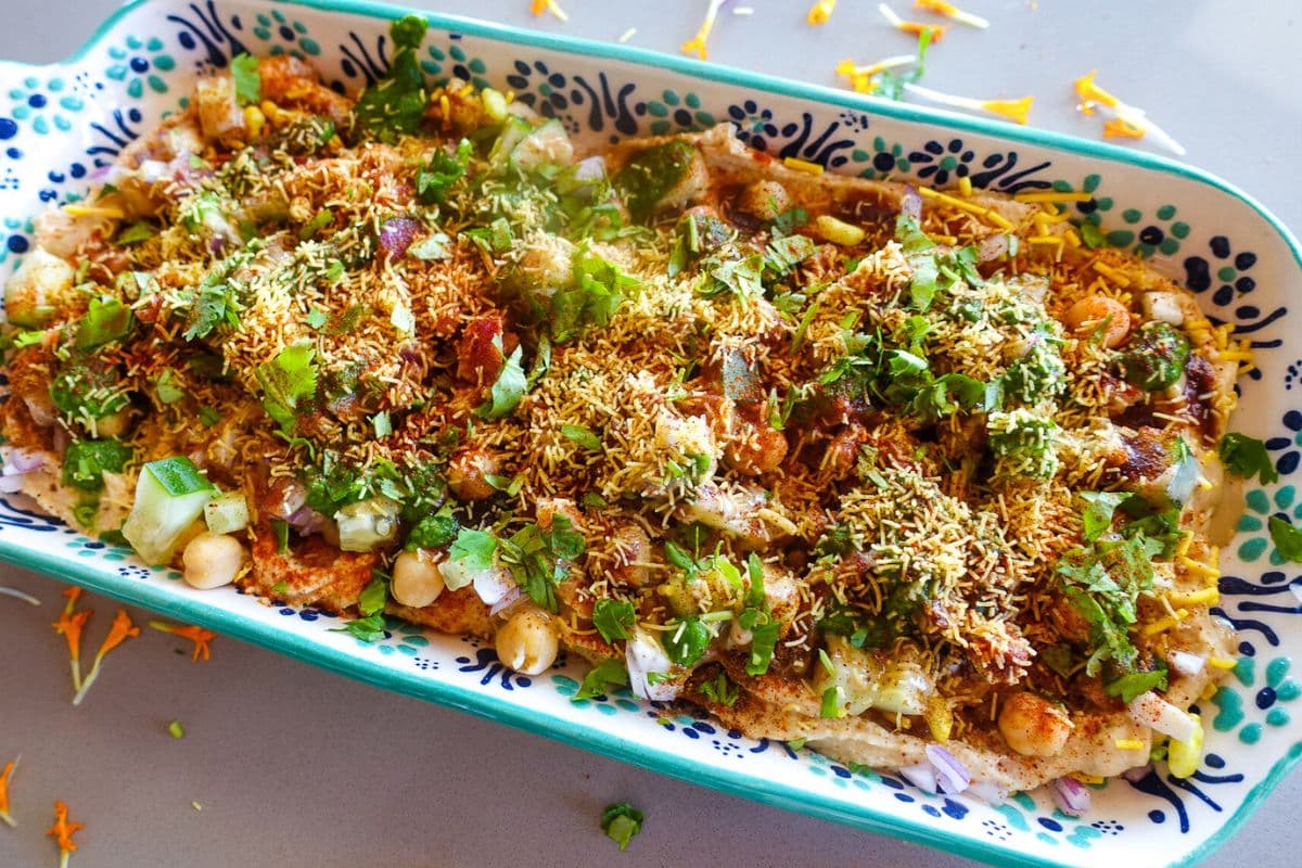 A rectangular dish filled with Hummus Chaat Dip topped with sev, cilantro, onions, chickpeas, and diced vegetables.