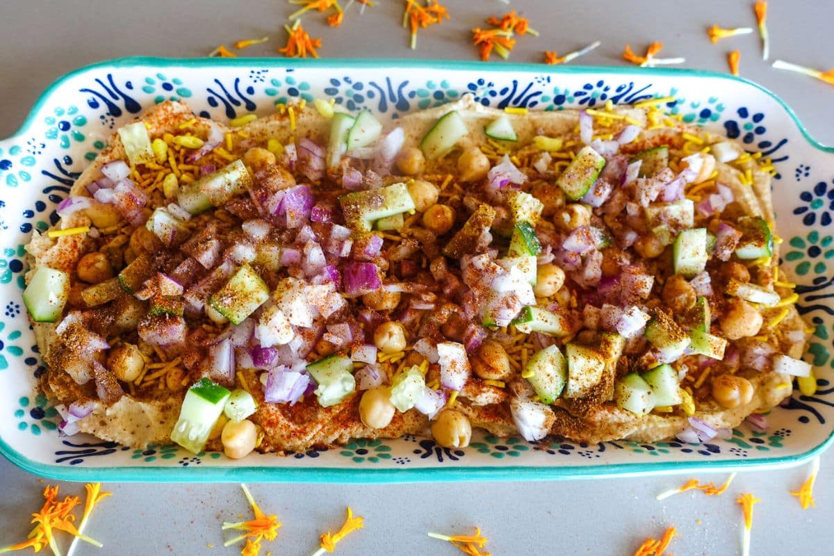 A rectangular dish with a layer of hummus, bhel mix, followed by chickpeas, chopped onions, and cucumber.