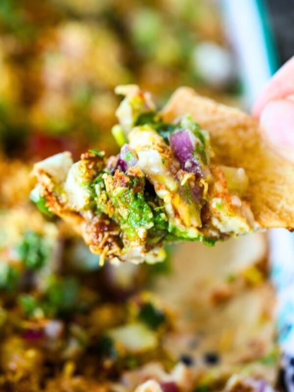 A hand holds a pita chip topped with Hummus Chaat Dip - a colorful mix of diced vegetables, herbs, and seasonings above a dish of dip.