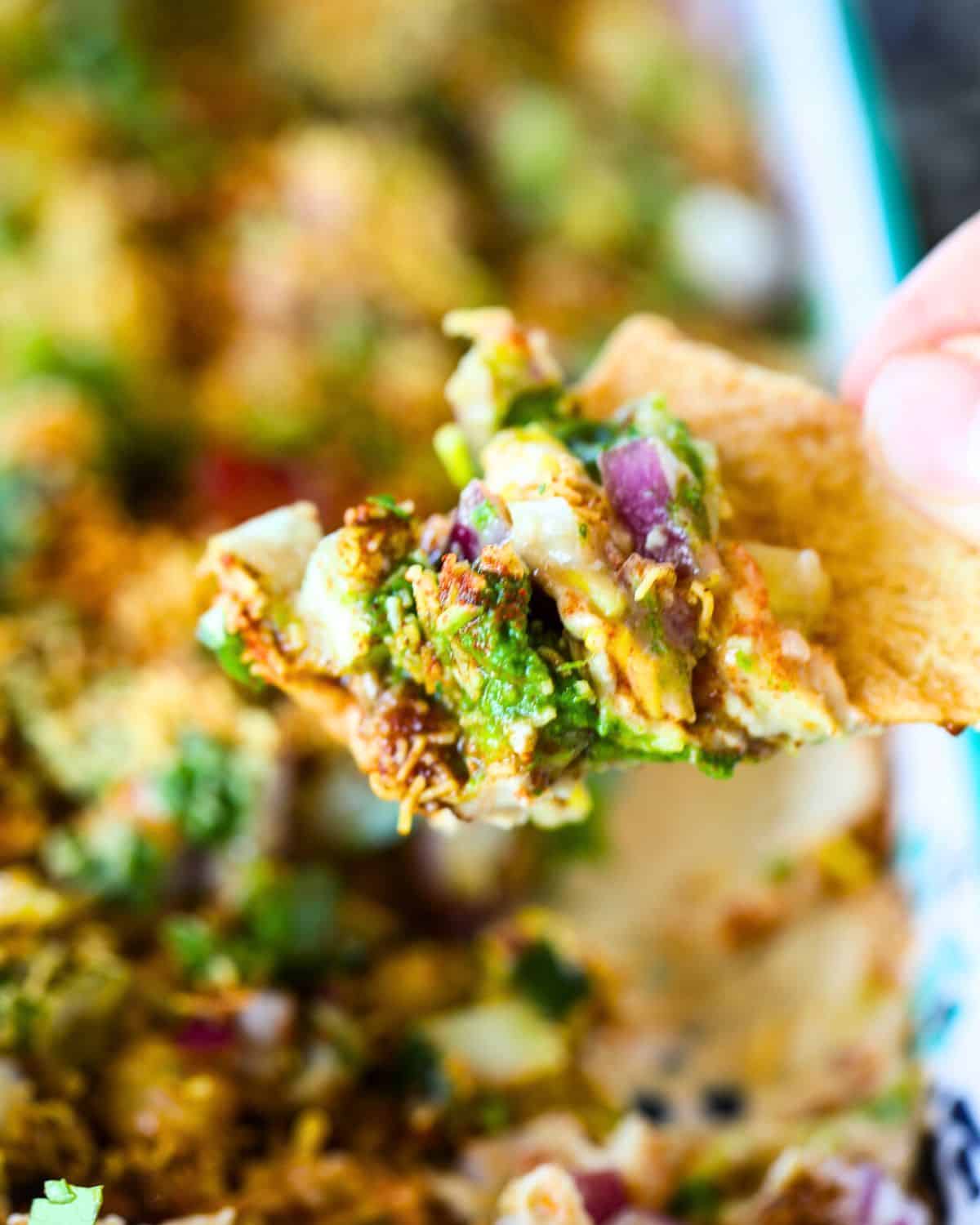 A hand holds a pita chip topped with Hummus Chaat Dip - a colorful mix of diced vegetables, herbs, and seasonings above a dish of dip.