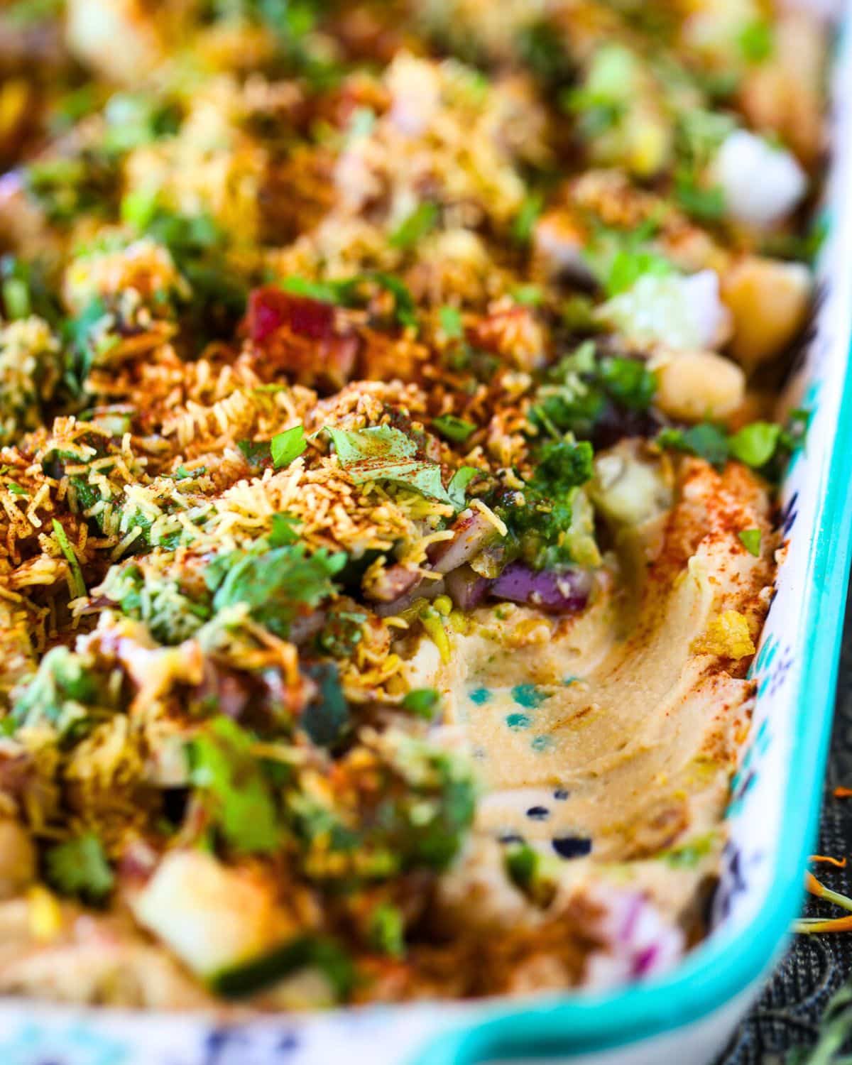 Close-up of Hummus Chaat Dip topped with chickpeas, chopped vegetables, herbs, spices, and sev in a rectangular serving tray.