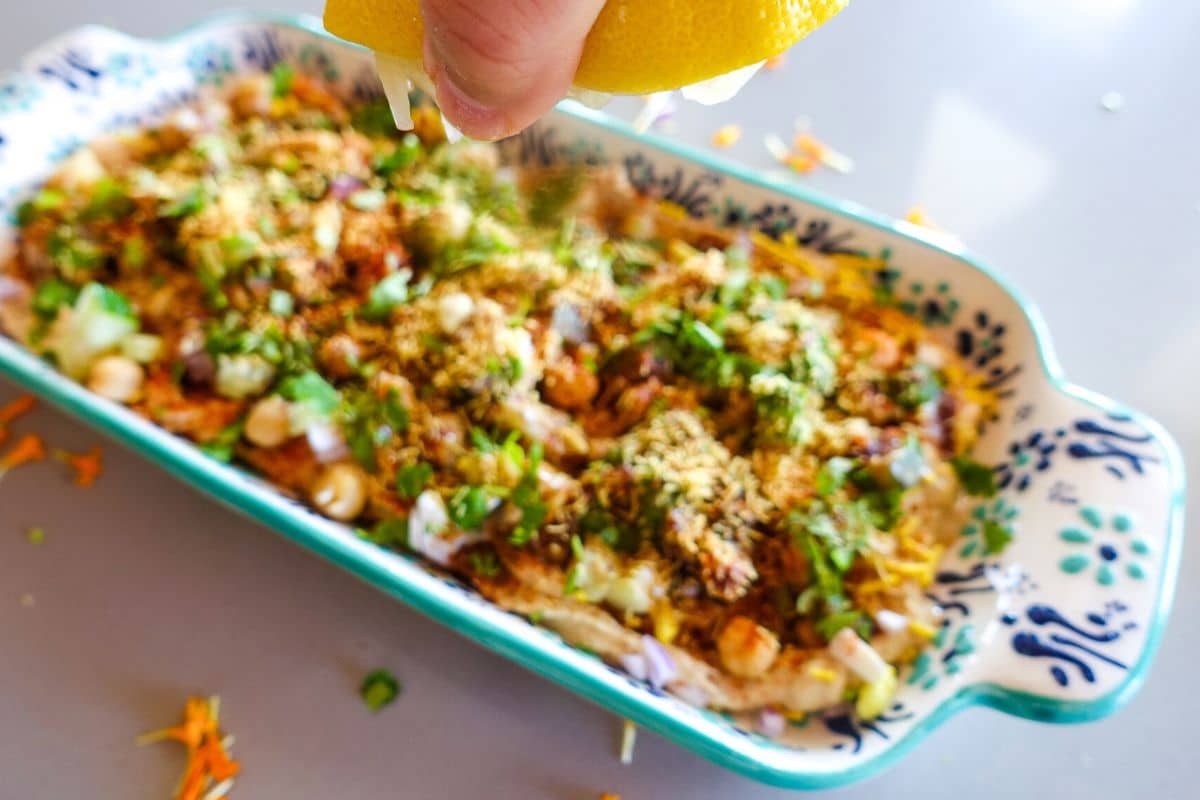 A hand squeezing fresh lemon over hummus chaat dip topped with herbs, chopped vegetables, and seasonings in a patterned ceramic tray.