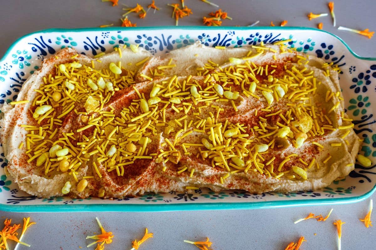 A rectangular dish of hummus topped with bhel mix and paprika with flower petals scattered on the dish and table.