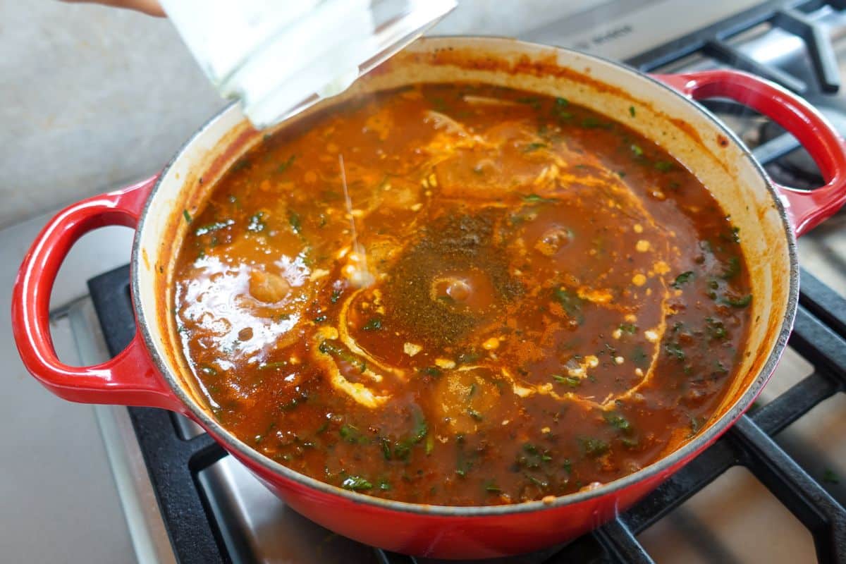 A hand pours cream into a red pot of tomato-based soup simmering on a stovetop, along with garam masala.