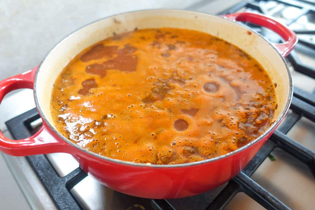A red pot filled with simmering stew or soup sits on a gas stove, with an orange and brown bubbling surface.