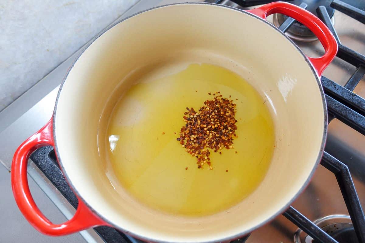 Red pepper flakes in oil are heating in a large red Dutch oven on a stovetop.