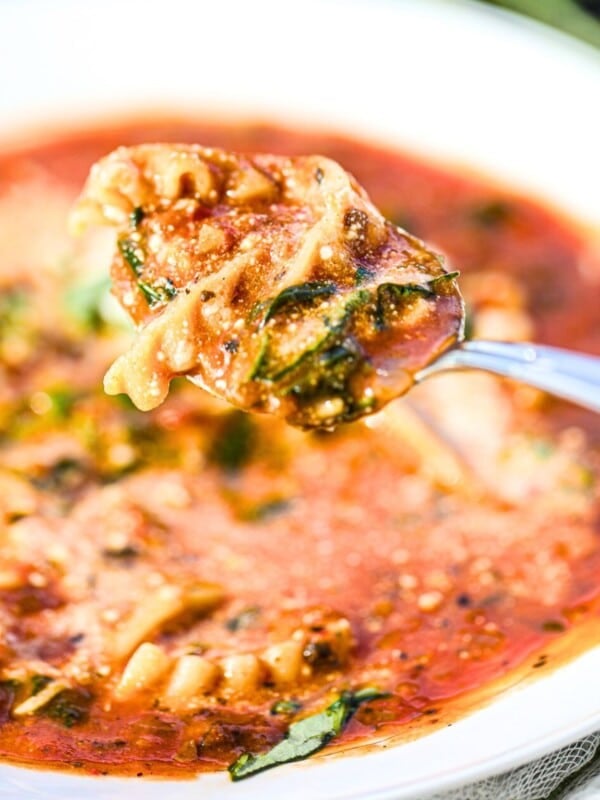 A spoonful of tomato-based soup with pasta, spinach, and herbs held above a white bowl filled with the same soup.