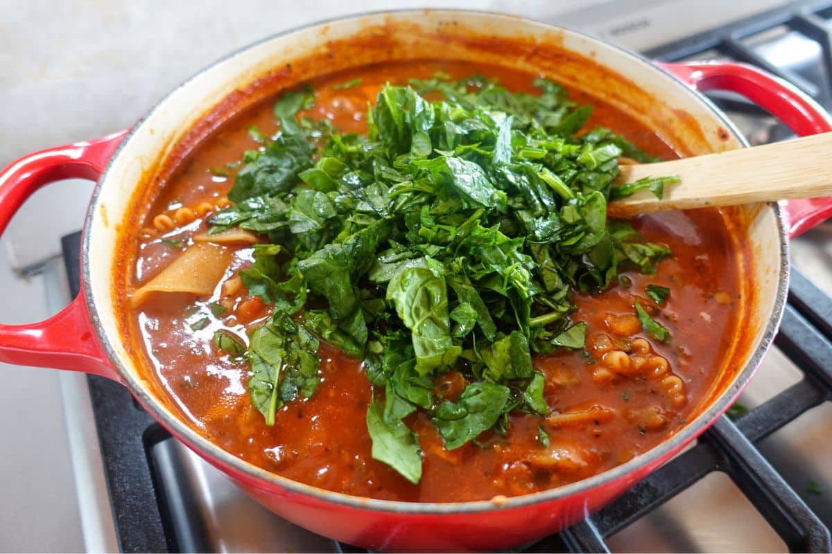 A red pot on a stove contains tomato-based soup with chopped spinach and lasagna noodles, being stirred with a wooden spoon.