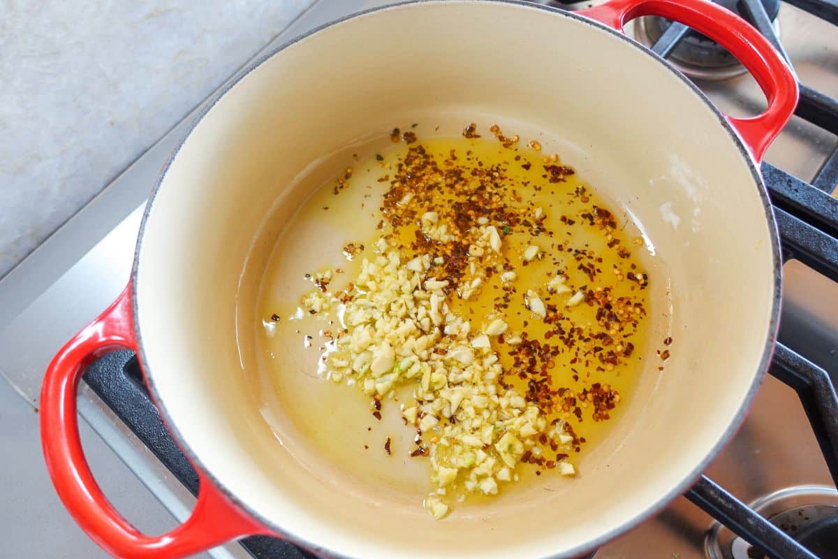 Chopped garlic and red pepper flakes saut&eacute; in olive oil in a red Dutch oven on a stovetop.