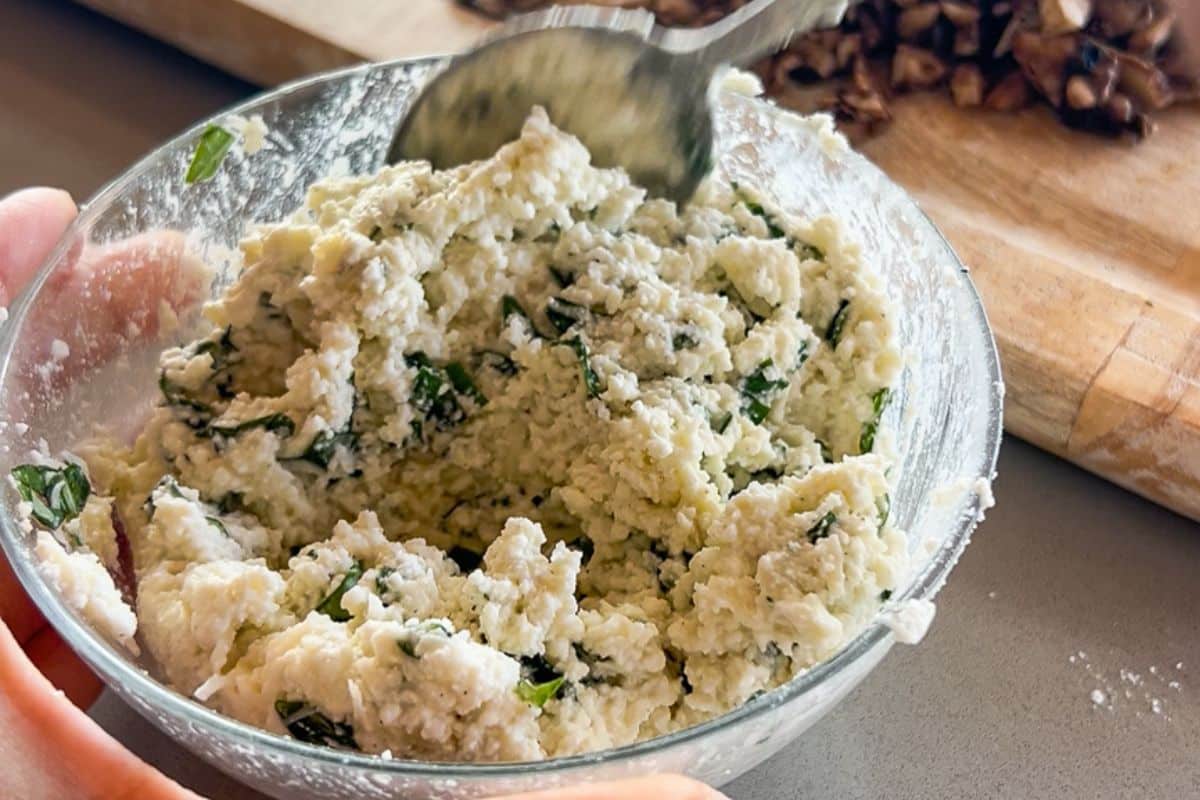 A hand holds a glass bowl of ricotta cheese mixture with herbs, being stirred with a spoon.