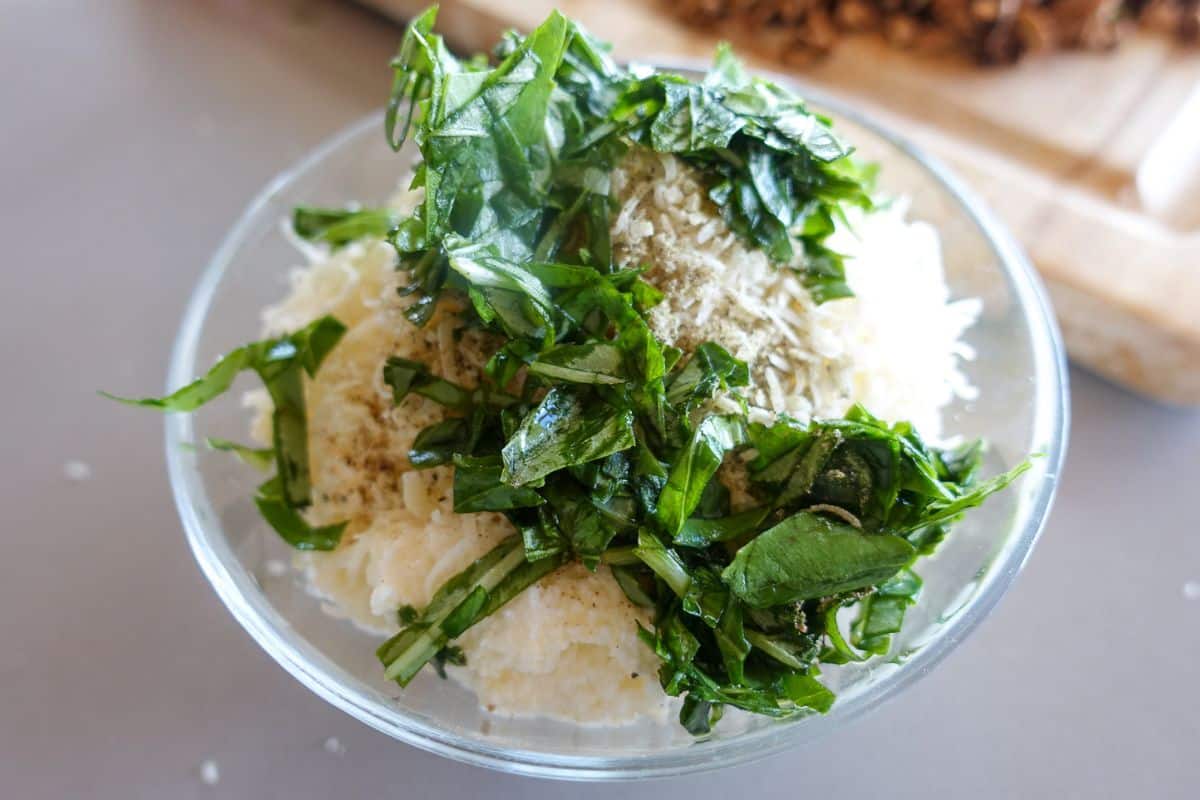 A glass bowl filled with grated parmesan, ricotta cheese, chopped fresh green herbs, and ground seasoning on a grey surface.