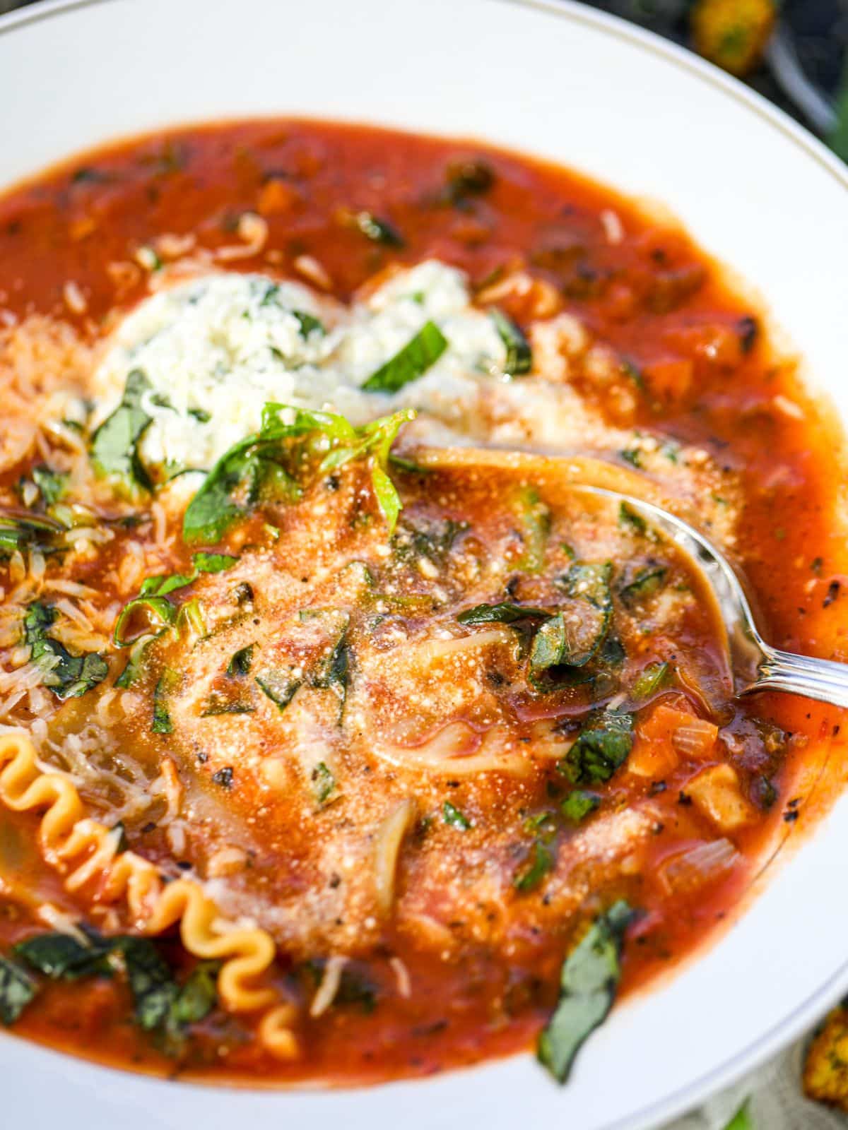 A bowl of lasagna soup topped with ricotta cheese, grated parmesan, and fresh basil, with a spoon resting inside.