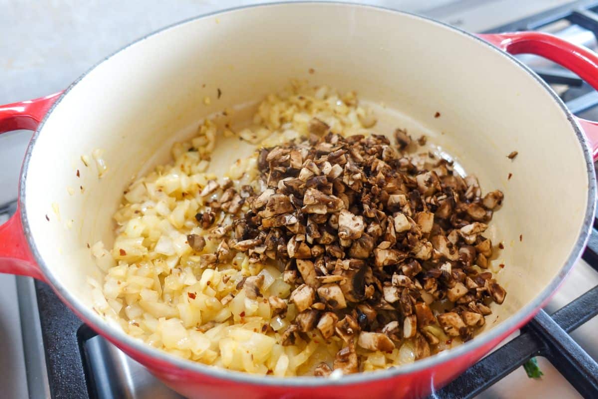 Chopped onions and mushrooms cooking in a red Dutch oven on a stovetop.