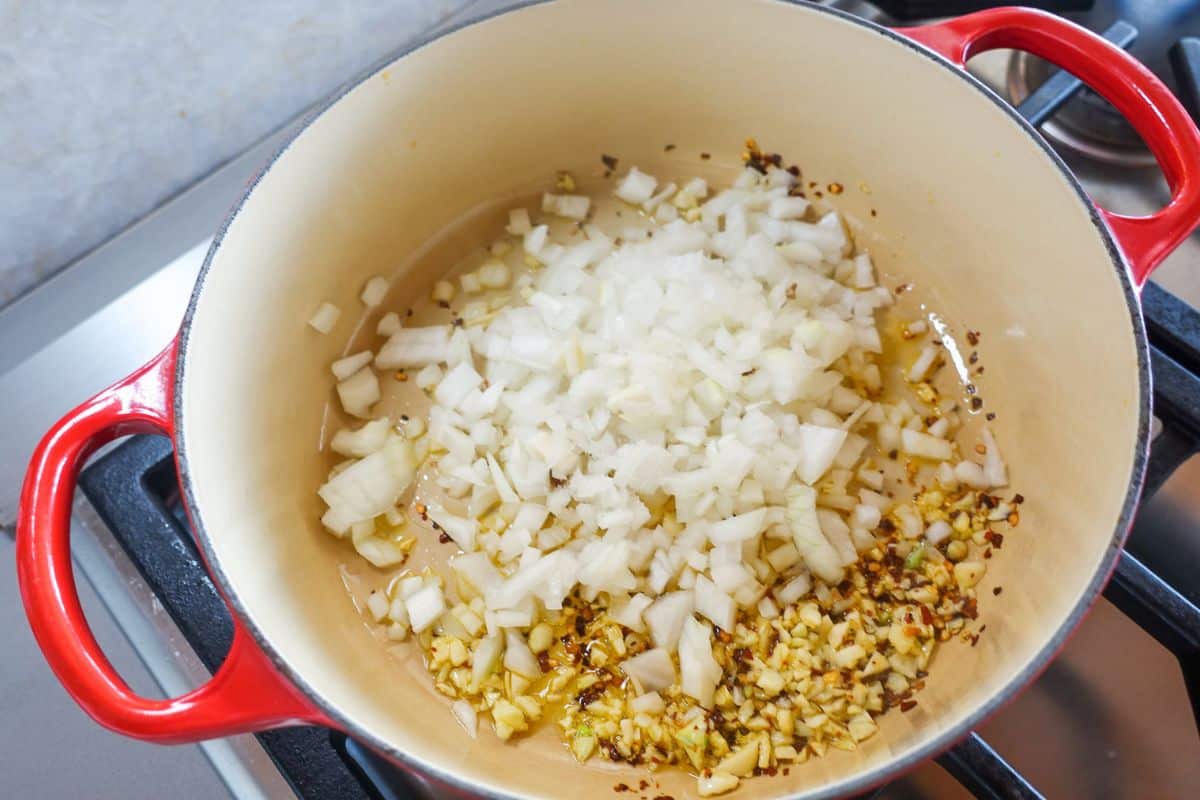 Chopped onions, garlic, and red pepper flakes cooking in a red Dutch oven on a stovetop.