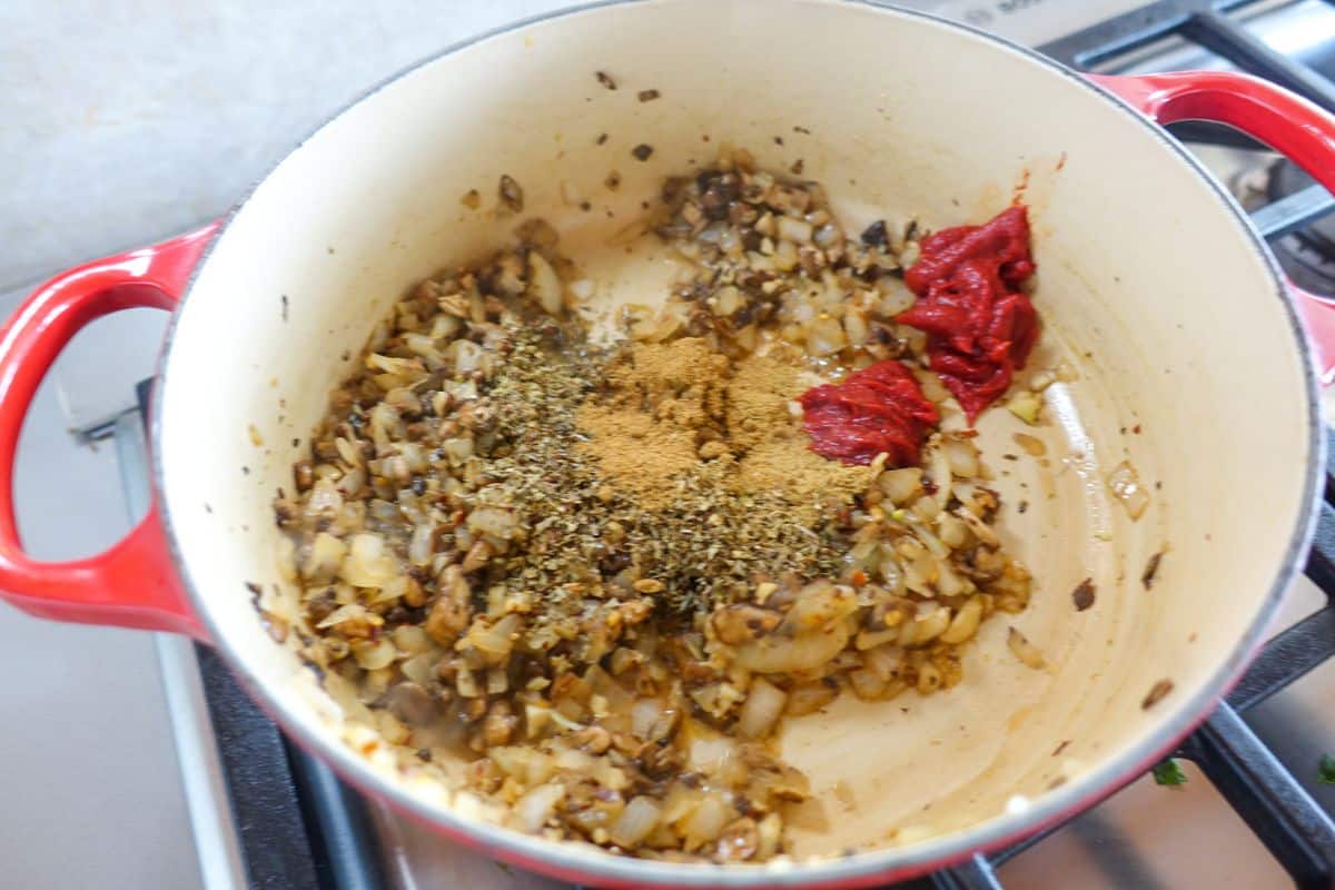 Chopped onions and mushrooms saut&eacute; in a red pot, with dried herbs, ground spices, and tomato paste added on top, on a stovetop.