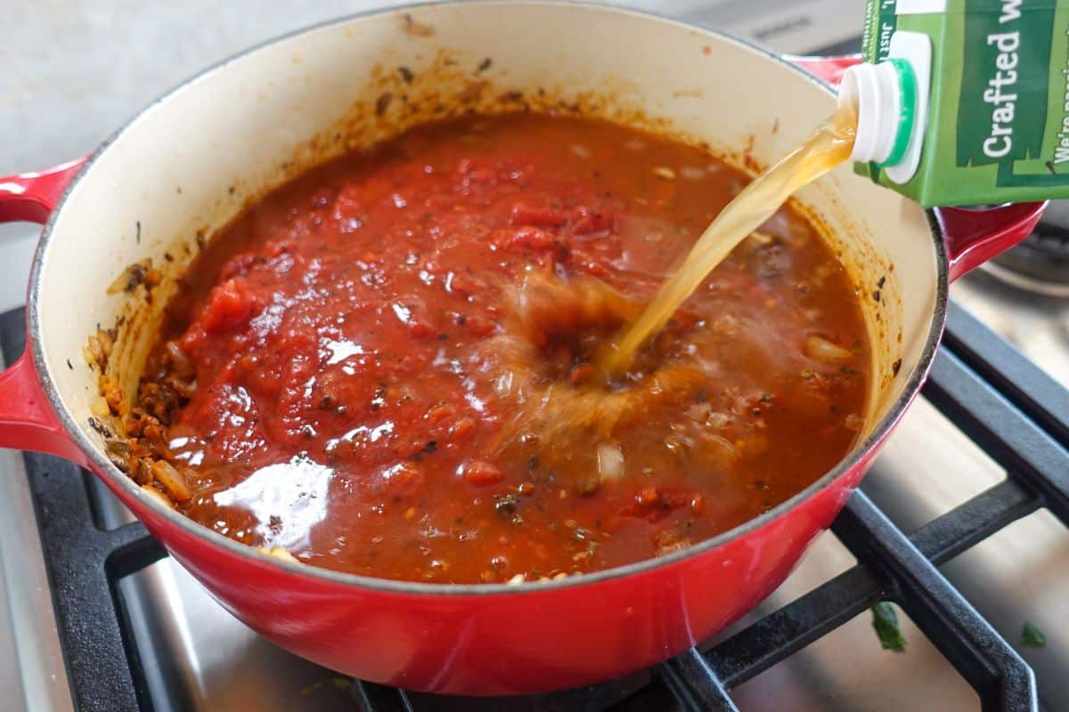 Vegetable broth is being poured from a carton into a red Dutch oven filled with crushed tomatoes on a stovetop.