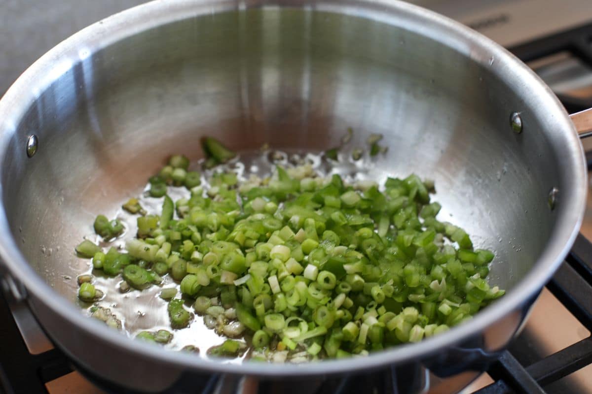 Chopped jalapenos and scallions cooking in oil in a stainless steel pan on a stove.
