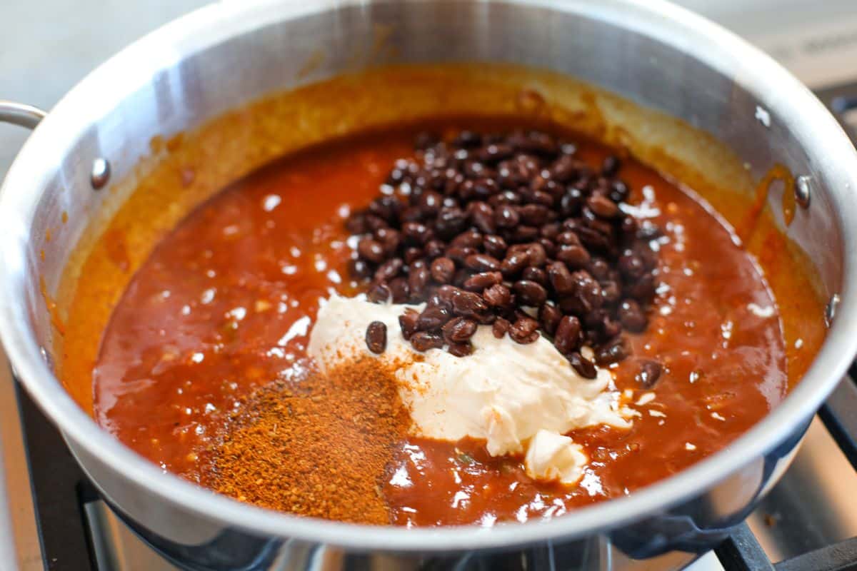 A pot of tomato-based sauce on a stove with black beans, taco seasoning, and sour cream added but not yet mixed.
